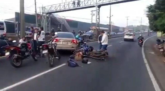 Road scene with multiple vehicles including cars and motorcycles some damaged and overturned a fallen motorcycle with helmet nearby people standing around possibly assisting or observing a pedestrian bridge in background and traffic congestion on the highway