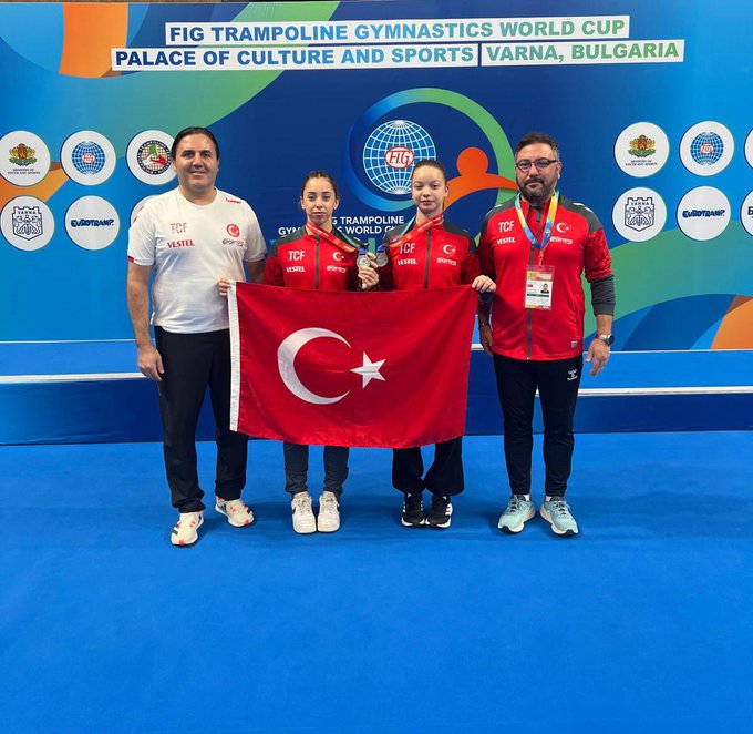 Four individuals stand on a blue gymnastics mat in front of a backdrop with FIG Trampoline Gymnastics World Cup and Palace of Culture and Sports Varna Bulgaria banners along with various flags and logos. A man in white shirt and two women in red Turkey uniforms hold a large Turkish flag. Another man in red shirt stands beside them. All wear medals around necks. The women appear as young athletes.
