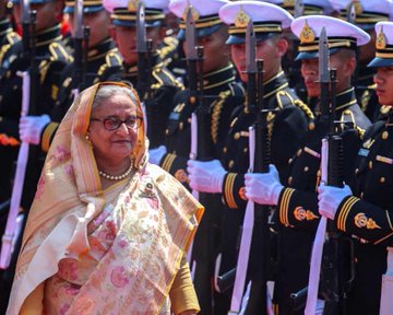 First image shows elderly woman in beige saree with floral patterns and glasses standing in front of line of uniformed men holding rifles wearing white gloves and navy caps with gold embroidery. Second image depicts street protest with thick white smoke billowing crowds of people some wearing helmets and backpacks near rickshaws and trees on urban road. Third image features same woman in light green saree sitting at wooden table with microphone gesturing with hand behind her with Bangladesh flag and potted plants.