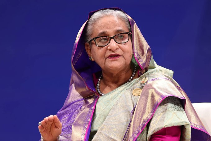 An elderly woman with gray hair covered by a purple-bordered headscarf wears thick-rimmed glasses and a green sari with intricate purple and gold patterns. She has a pearl necklace and a badge on her chest. Her expression is serious as she gestures with her hand. The background is solid blue.