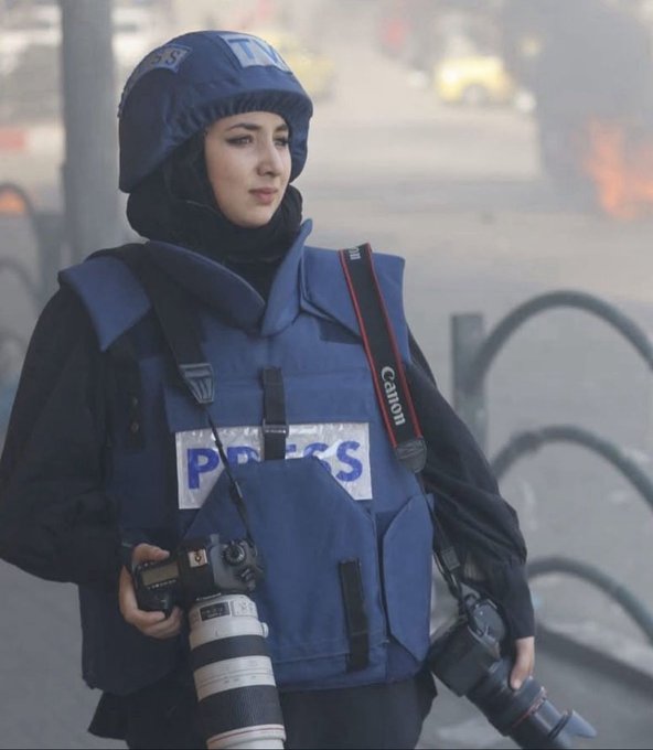 A woman wearing a blue hijab and protective vest labeled PRESS stands in a street scene with smoke and fire in the background holding a camera with a long lens and strap over her shoulder next to a yellow vehicle and metal barriers.
