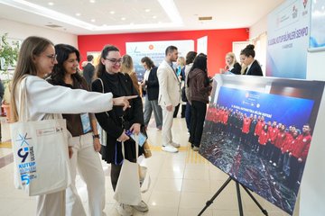 First image shows a speaker on stage presenting on Arctic topics with a large screen displaying a world map highlighting polar regions blue text on Arctic engineering and polar bears in an auditorium with audience seated and a banner for 9th National Polar Sciences Symposium. Second image depicts a group of young women and men in casual and formal attire pointing at a display screen showing a team in red uniforms possibly researchers with a banner for Polar Symposium and tote bags nearby in a red-walled room. Third image features a large group of young adults in black and white clothing posing for a group photo in front of large blue letters spelling 5.K.KUTUPUSENLİĞİ with trees tents and buildings in the background on a paved area. Fourth image captures an audience seated in a large auditorium watching a presentation on a screen with polar map projections during the Polar Sciences Symposium with banners and chairs arranged formally.