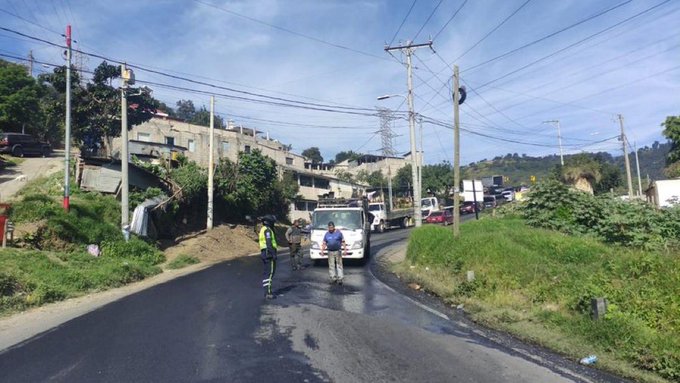 Road scene at kilometer 26 on route CA-9 with pothole repair work in progress a white vehicle and workers in high-visibility vests stand near the wet asphalt surface surrounded by utility poles overhead wires and nearby buildings and vegetation reversible lanes are coordinated in the area