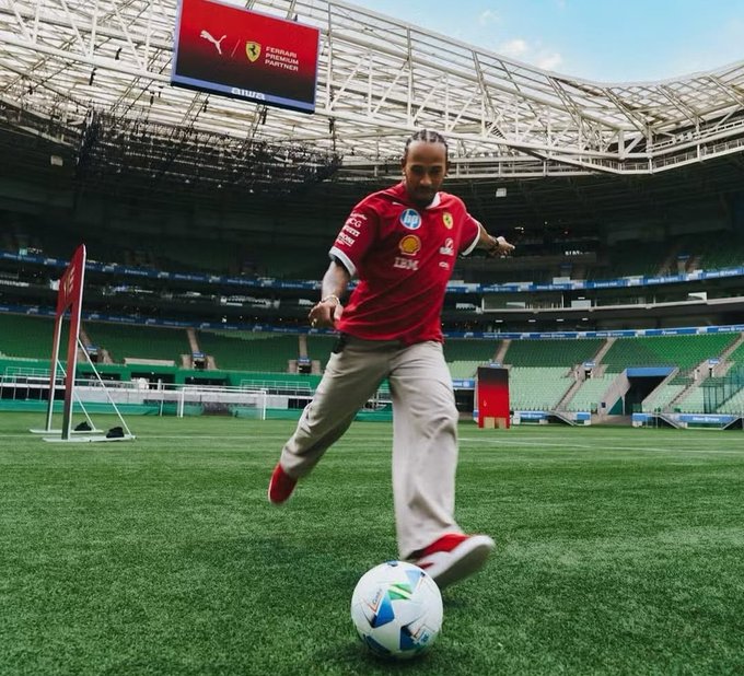 Lewis Hamilton in red Ferrari Puma jersey with sponsor logos and beige pants kicks blue and white soccer ball on green field in stadium. Red Puma Ferrari screen displays on structure above. Green stadium seating and banners visible in background. Lewis Hamilton has dreadlocks and is mid-action with one leg raised.