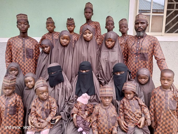 Group photo of approximately 20 people including men women and children standing together outdoors against a green wall and window. Most wear traditional brown and patterned robes abayas and head coverings with some men in embroidered caps and attire. Several young children visible some holding babies all dressed similarly in cultural garments suggesting a family gathering.