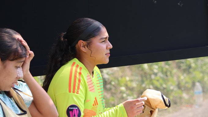 Two female soccer players from the Mexican Under-17 team wear yellow Adidas jerseys with black stripes and sponsor logos including VW. One player with long dark hair in a ponytail looks focused while holding a jersey, standing near a black background with green foliage visible. The other player with curly hair touches her head, both on a field setting.