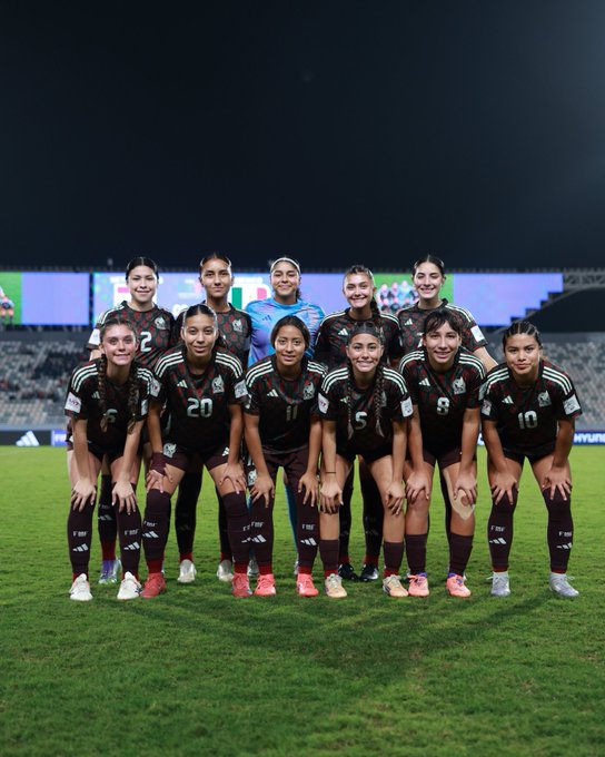 Group of eleven women soccer players from the Mexican national under 17 team standing and kneeling on a grassy field at night in a stadium. They wear dark green jerseys with white accents and numbers like 20 and 11, dark shorts, and white socks with green tops. The background shows a lit stadium with green field and stands. Adidas logos are visible on jerseys.