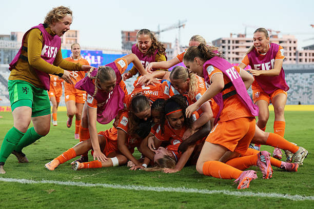 Group of female soccer players in orange jerseys and pink vests celebrating by piling on top of each other on green grass field with referee in green uniform nearby stadium seating and construction cranes in background under clear sky