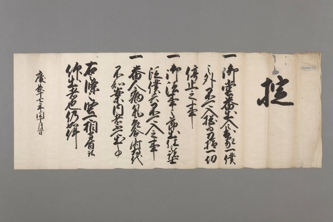 Ancient Japanese scroll on aged paper with black ink calligraphy in vertical columns, featuring formal kanji characters outlining rules in a historical document style, displayed in a museum setting with neutral background.