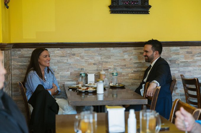 First image shows a man in a dark suit and tie standing next to a woman in a light blue button-up shirt and white pants both smiling in a restaurant with yellow walls a framed Buddha statue on the wall potted plants wooden tables with plates and chairs in the background. Second image displays a wooden table with multiple metal thali plates containing yellow curries red sauces vegetable dishes like pakoras and dumplings white rice naan bread glasses of tea and a salt shaker. Third image depicts the same man and woman seated at a table laughing with plates of food water bottles and glasses in front of them against a yellow wall with decorative elements and chairs.