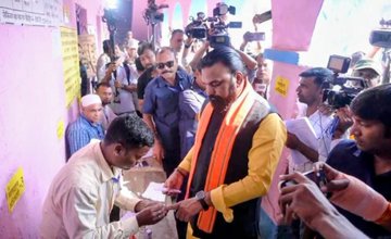First image depicts a man with long hair and beard wearing yellow kurta black vest saffron scarf holding up index finger in voting gesture wristwatch visible plain background. Second image shows the same man in yellow attire saffron scarf exchanging something with another man while surrounded by media personnel holding cameras and microphones in pink-walled room with posters. Third image portrays a crowd of men and women in colorful saris and kurtas gathered outside a blue and pink polling station building with arched entrances sign reading voting center rural setting with trees.