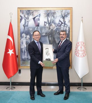 First image shows two men in dark suits standing side by side in front of Turkish and white flags with a large framed painting of people on a wall behind them in an official room with blue carpet. Second image depicts the same two men holding a framed document or plaque with calligraphy between the flags and painting in similar setting. Third image features a group of men and women in formal attire seated around a wooden table in a room with Turkish flag Chinese flag and ornate decor including curtains and a painting on the wall.