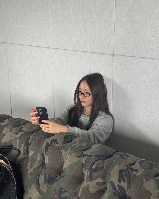 Woman with long dark hair wearing glasses and gray sweater sits on camouflage-patterned couch holding red-cased smartphone. Man with headphones sits in checkered chair in brick-walled studio with white wall, microphone stands, speakers, turntable, and window showing trees. Woman with long hair wearing headphones and striped shirt with shorts stands in dark room with foam panels, mixing console, and equipment.