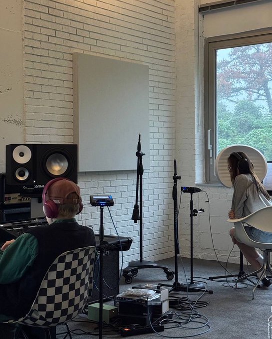 Woman with long dark hair wearing glasses and gray sweater sits on camouflage-patterned couch holding red-cased smartphone. Man with headphones sits in checkered chair in brick-walled studio with white wall, microphone stands, speakers, turntable, and window showing trees. Woman with long hair wearing headphones and striped shirt with shorts stands in dark room with foam panels, mixing console, and equipment.