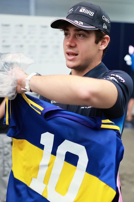 A young man with short dark hair wearing a black cap with sponsor logos and a dark short-sleeved shirt stands indoors against a plain white wall. He holds a blue and yellow vertically striped soccer jersey displaying the number 10 on the back with his hands gloved in clear plastic. His expression is smiling and engaged.