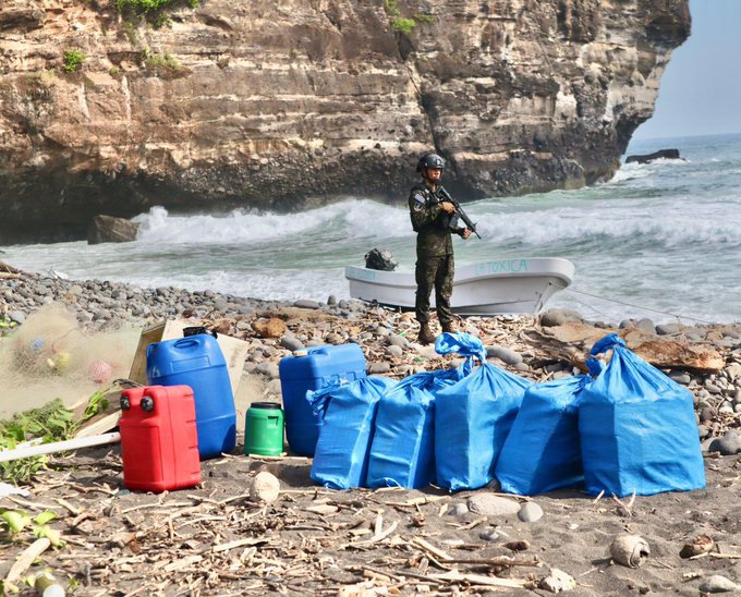 A member of the armed forces in camouflage uniform and helmet stands holding a rifle next to a small white motorboat on a sandy beach with scattered rocks and vegetation. Several blue plastic bags and containers possibly holding drugs are placed on the ground nearby along with red and green fuel cans and other supplies. In the background rocky cliffs rise above the ocean with waves crashing and a hazy sky.