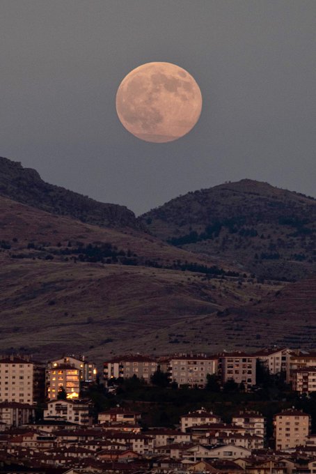 A large full moon glows orange against a twilight sky above rugged brown hills covered in sparse vegetation. Below the hills, a cluster of white multi-story apartment buildings with red-tiled roofs lines the base, some illuminated by warm lights. The scene captures the Super Moon in its closest approach to Earth, appearing larger and brighter than usual. Another similar image shows the pinkish moon rising over the same landscape with buildings. A book cover displays a cosmic scene with stars and galaxies in orange and blue hues, titled Gökyüzünü Tanıyalım by M. Emin Özsah, published by TÜBİTAK. An open book page features diagrams of the moon with craters, phases, and textual explanations in Turkish about lunar features and exploration.