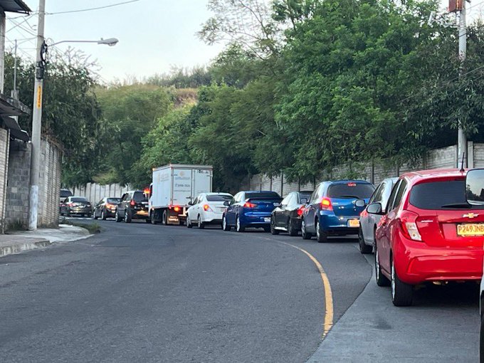 First image displays a line of vehicles including cars trucks and vans stopped on a curved road lined with trees walls and utility poles under overcast sky with some emergency lights visible. Second image shows similar congestion with additional traffic cones placed on the road near a silver car and more vehicles extending into the distance along the tree-lined street with a hill in background.