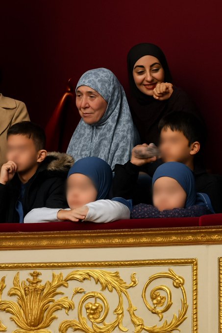 Group of four blurred-faced children aged around 10-11 wearing blue veils, long blue garments, and dark clothing sit on red velvet seats in a grand hemicycle with ornate gold decorations on white panels and red walls behind them, accompanied by two adult women in headscarves and coats.