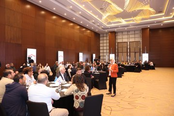 First image displays a young girl in a blue shirt standing on green grass with arms raised happily in front of a school building under a blue sky with bubbles, featuring Turkish flag shield emblem labeled Tertemiz Yarışması Sosyal Sorumluluk Yarışması Başlıyor with text on cleaning and hygiene project details, Son Başvuru 10 Kasım 2023, QR code, and logos of MEB and OPET. Second image shows a large group of adults in business attire standing together on a patterned floor in a conference hall with decorative walls and screens displaying Turkish flags, OPET logo, and event branding. Third image depicts a woman in orange top and black pants speaking at a podium in a modern room with wooden walls, hanging lights, multiple screens showing project visuals, and audience seated at round tables with drinks and notebooks. Fourth image illustrates a man in suit speaking at a podium in a similar conference setting with wooden decor, screens displaying shields and OPET logo, audience at tables including diverse group of men and women in formal wear.