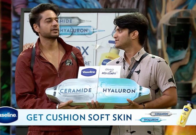 Two young adult men stand on a colorful Bigg Boss show set with graffiti walls and sponsor banners. The man on the left wears a red zip-up jacket over a graphic tee and holds a blue Vaseline Hyaluron bottle. The man on the right wears a light beige short-sleeve shirt and holds a white Vaseline Ceramide bottle. Promotional text reads Get Cushion Soft Skin with Vaseline logos visible.