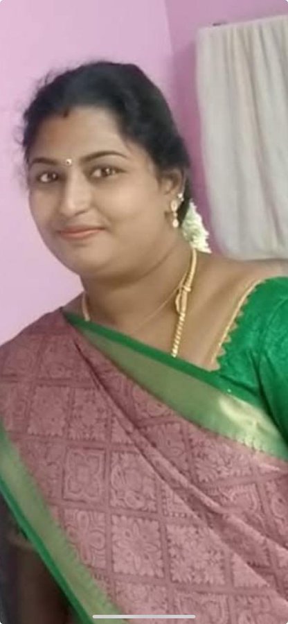 A woman with long dark hair braided and adorned with jasmine flowers wears a green-bordered maroon saree over a green blouse and gold jewelry including necklace earrings and bangles stands against a pink wall with a white curtain in the background