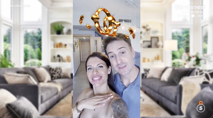 A smiling woman with long dark hair and a man with short hair pose closely together in a modern living room featuring large windows, green plants, bookshelves, and gray sofas. Golden ring emojis float above their heads. The setting includes decorative elements like vases and cushions.