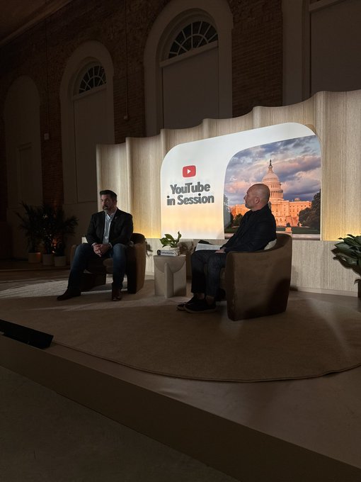 Two men are seated on a modern stage with light wood paneling and potted plants. The man on the left wears a dark suit and sits in a white chair, while the man on the right wears a dark sweater and sits in a brown armchair. Behind them is a large curved backdrop with the YouTube logo and an image of the US Capitol building under a blue sky. The setting appears to be an indoor event space with arched windows and brick walls in the background.