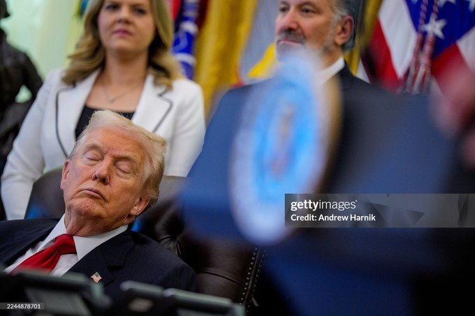 An image shows Donald Trump sitting in a leather chair with eyes closed and mouth slightly open appearing asleep wearing a dark suit red tie and American flag pin in a formal room with American and other flags in background a woman in white blouse on left a man in suit on right and a blue round object in foreground credit to Andrew Harnik via Getty Images.