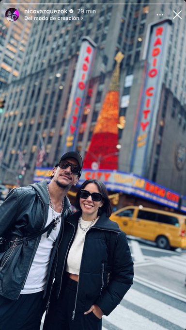 A man and woman stand closely together smiling at the camera in front of a large neon-lit building with Radio City signage and Christmas decorations. The man wears a black leather jacket, white shirt, jeans, and sunglasses, with a chain necklace. The woman wears a white puffer jacket, black pants, sunglasses, and a necklace. A yellow taxi is parked nearby on the street.