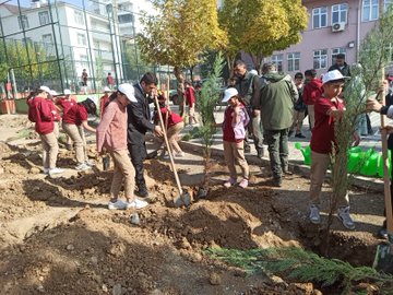 First image shows several suited men and children in red uniforms and white hats planting and watering young saplings in dug soil near a chain-link fence with apartment buildings and trees in the background under a clear sky. Second image depicts children in red hoodies and white hats kneeling to plant a sapling next to a police vehicle on a paved area surrounded by fences and buildings. Third image captures a group of children in uniforms and adults including suited individuals digging and planting multiple saplings in a row along a fence with green watering cans nearby and buildings in view. Fourth image features a group of children in red uniforms and adults standing near planted saplings and dug soil patches in an open area with fences, trees, and apartment buildings.