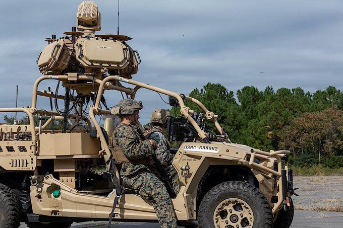 A tan military vehicle resembling a Polaris MRZR with a mounted sensor array and antenna on top sits on a dirt path amid pine trees under a cloudy sky. Two soldiers in camouflage uniforms and helmets are positioned in the vehicle, one driving and the other in the passenger seat. USMC markings are visible on the vehicle and soldiers gear.