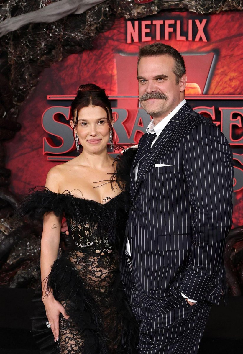 Millie Bobby Brown stands beside David Harbour on a red carpet, both smiling at the camera. Brown wears a black lace gown with off-shoulder black feather accents and a high slit, her hair in an updo. Harbour is dressed in a dark pinstripe suit with a white shirt and tie, sporting a mustache. Behind them is a promotional backdrop for Netflix Stranger Things featuring the title in bold letters, rocky terrain, red glow, and hanging cobwebs.