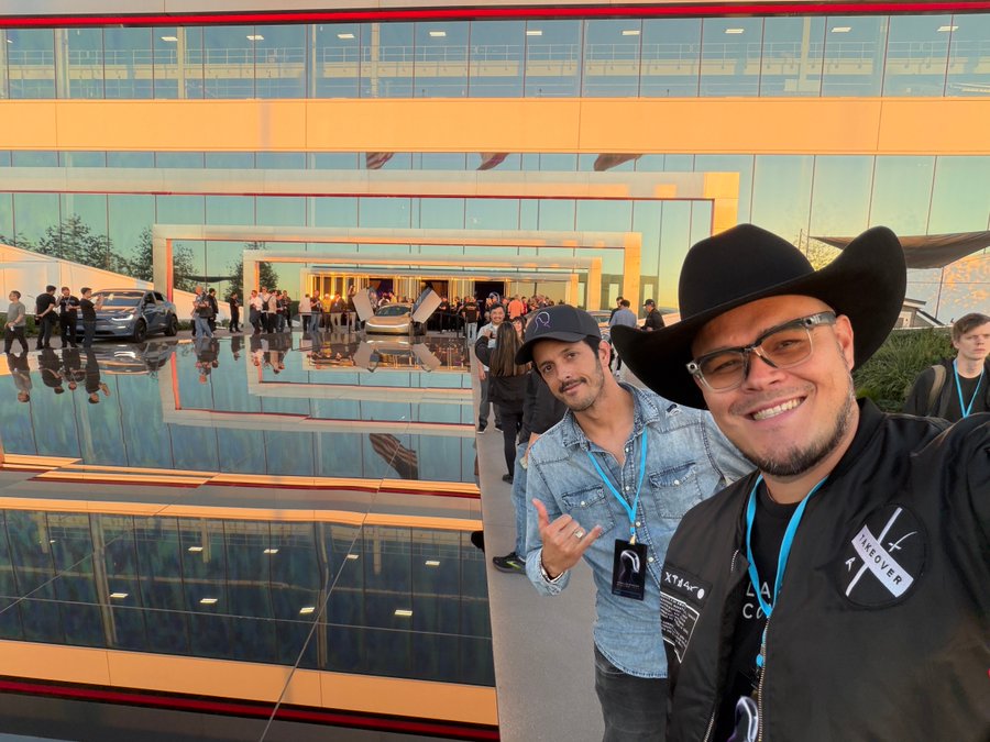 Several people stand on a reflective outdoor platform surrounded by modern glass architecture with orange sunset hues. In the foreground a man with glasses and a black jacket wearing a lanyard makes a peace sign while another man in a cowboy hat and sunglasses smiles beside him. In the background multiple white Tesla Cybertrucks are parked near a yellow building with people gathered around. The scene includes event attendees in casual attire and lanyards.