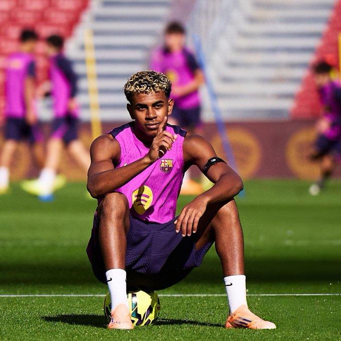 Young male athlete with curly blonde hair and light skin wearing sleeveless pink FC Barcelona training top purple shorts white armband white socks and orange Adidas cleats sits on green soccer field with yellow-green soccer ball positioned under his right foot hand touching chin in thoughtful pose blurred teammates in matching pink uniforms train in background red stadium seating and green pitch visible.