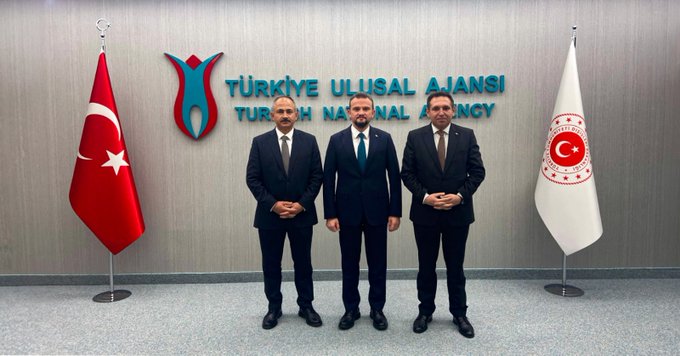 Three men in dark suits and ties stand side by side in front of a wall with the Turkish Ulusal Ajans logo and Turkish flags on poles flanking them the central man has a mustache and the group poses formally in an office setting with a hanging banner displaying Türkiye Ulusal Ajansı text.