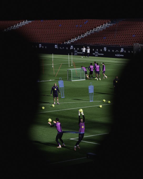Green soccer field in a stadium with red tiered seats and black barriers in the background. Training equipment including orange cones, blue and white goalposts, and white poles scattered across the pitch. Several players in purple jerseys and shorts engaged in drills, one player in purple raising arms near the goal, another in purple catching a yellow soccer ball mid-air near the blue goal net. Stadium lighting creates a focused glow on the field.