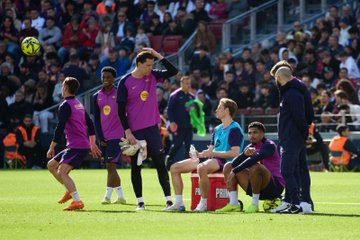First image shows a male soccer player in purple kit with yellow accents jumping to head a yellow soccer ball near white goalposts on green field in stadium filled with blurred spectators in stands, advertising banners visible at bottom. Second image depicts group of male athletes in purple and blue training uniforms standing and sitting on green pitch with yellow soccer ball in air, water cooler nearby, stadium seating in background. Third image features young male player with curly hair in purple sleeveless top facing coach in dark jacket and yellow text on back near green field with yellow balls scattered around, stadium stands behind. Fourth image captures coach in dark jacket speaking to young male player in purple tank top and shorts under spraying water on green turf with yellow soccer ball at feet, goal net and field markings visible.