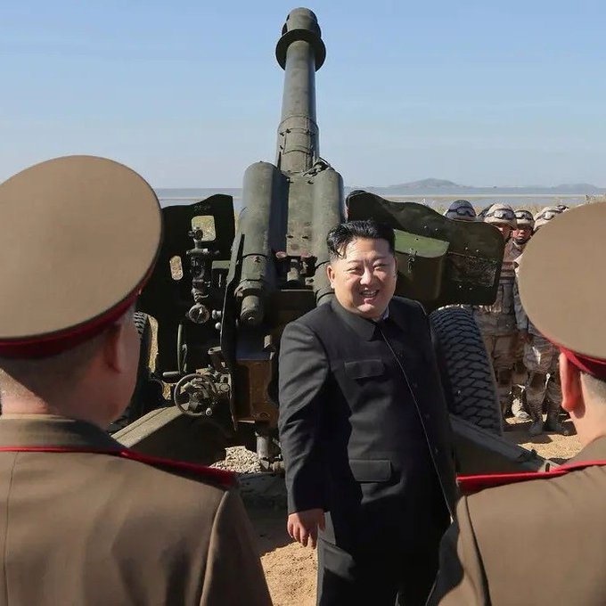 Kim Jong-un stands smiling in a black suit and red tie beside a large green artillery cannon on a sandy desert terrain under a clear blue sky. Several North Korean soldiers in tan uniforms and caps surround him, some facing the cannon. In the background, additional soldiers in camouflage gear stand near military vehicles on the open field with distant hills visible.