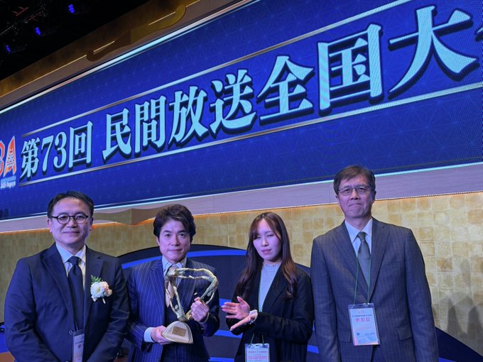 Four people stand on a stage with a large blue banner displaying Chinese characters and the text 73民放協 in the background. The individuals are dressed in formal suits and dresses, two men and one woman holding a trophy shaped like a vase with a ribbon, another woman gesturing with her hand. Name tags are visible on their clothing, and the setting appears to be an indoor convention hall with gold patterned walls.
