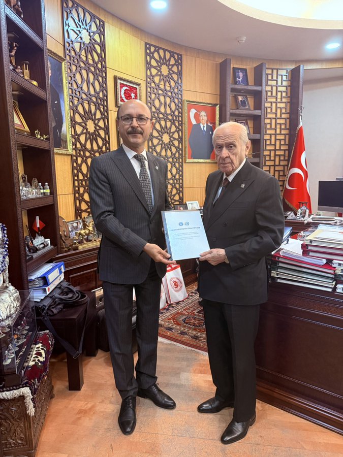 Two men in dark suits stand in a wooden-paneled office with Turkish flags and portraits on walls, one holding a white certificate folder labeled in Turkish, bookshelves and decorative items visible around a desk.