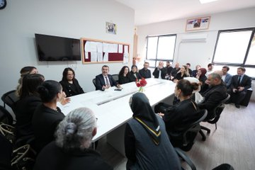 First image shows a man in a suit with tie embracing two young boys wearing backpacks and jackets one blue with a logo outside a building with brick walls and windows. Second image depicts the same man in suit smiling and touching cheeks of a group of young girls and boys in red uniforms raising hands in a school hallway with green exit signs and wooden beams. Third image captures the man seated at a long white table in a meeting room with women in black attire some wearing headscarves flowers on table and wall decorations including framed photos and a TV screen. Fourth image features a group of adults in formal suits and dresses standing on steps in front of a yellow school building entrance labeled Bursa Karacabey 14 Eylul Ortaokulu with trees and blue sign.