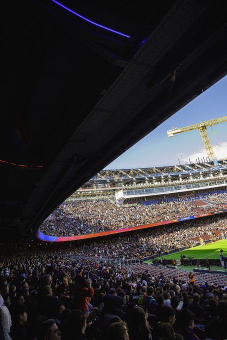 A wide-angle view from the upper seating area of a large soccer stadium shows ongoing construction with a prominent yellow crane extending into the clear blue sky. Crowds of spectators fill the blue and red seats below creating a vibrant sea of people in casual clothing. The green field is visible in the center surrounded by the curved stadium architecture under bright daylight.