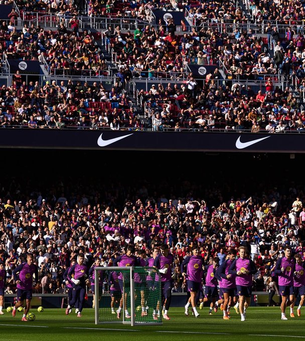 Crowded stadium stands filled with fans in various colors cheering, green soccer field with goal net and posts, Barcelona players in purple jerseys and shorts warming up by running and stretching near the goal area, blue team players in dark uniforms visible, Nike swoosh logos on black stadium signage above seating sections.