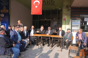 First image shows a group of men in suits and casual clothes walking on a street near a red Anafarta market building with green awnings and shops, including a boy in a black hoodie among them, under autumn trees. Second image depicts several men in suits and traditional attire seated around wooden tables outdoors under a Turkish flag, engaged in discussion at what appears to be a meeting. Third image features men in suits standing near hanging colorful childrens clothing like pink puffy jackets and dresses on racks, with a young girl in a yellow top beside them in an outdoor market setting with a toy horse. Fourth image shows a man in a suit at a blue tented stall with women in headscarves serving jars of liquids and food items like bottles and cans to visitors in a lively bazaar environment.
