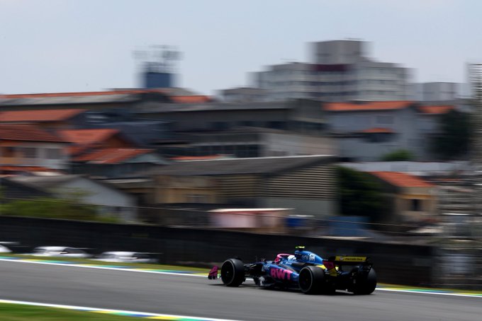 Blue and pink Alpine Formula 1 car races on a track during a session with white barriers and green astroturf on the sides surrounded by blurred urban buildings with red roofs and a tall structure in the background under overcast skies