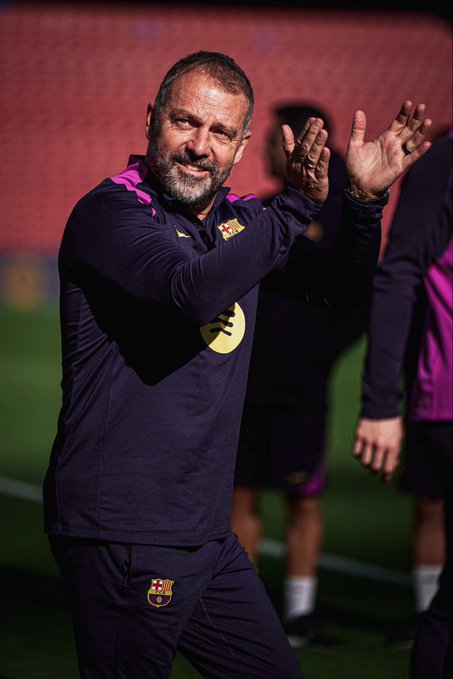 First image shows a middle-aged man with short gray hair and beard, identified as Hansi Flick, clapping enthusiastically with both hands raised, wearing a long-sleeve navy tracksuit with purple accents and yellow Nike swoosh, Barcelona crest on left leg, standing on green grass field near brick wall with purple trim, two blurred figures in similar attire behind him, stadium seating visible in background. Second image depicts the same man from side angle, smiling faintly, hands at sides, in identical tracksuit, positioned on field with red stadium stands and green grass extending behind, no other distinct figures prominent.