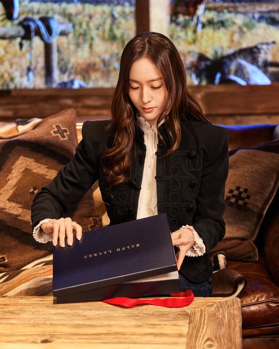 First image shows a woman with long black hair wearing a brown knit top holding up a white cable-knit sweater with a small logo near a wooden table with an open black box and red ribbon in a room with a stone fireplace and wooden logs. Second image depicts the same woman in a black jacket over a white collared shirt opening a navy blue box with red ribbon on a wooden table beside beige cushions and a window with greenery outside. Third image features a young man with short black hair in a patterned knit cardigan over a denim shirt sitting on a brown leather chair with legs crossed near a wooden table holding a navy blue box with red ribbon and patterned cushions in a room with large windows showing trees.