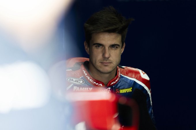 A young man with short brown hair and a focused expression wears red white and blue racing leathers featuring sponsor logos including Honda Arai and American Racing on the chest and arms stands in a dimly lit indoor setting with a blurred background suggesting a garage or team area.