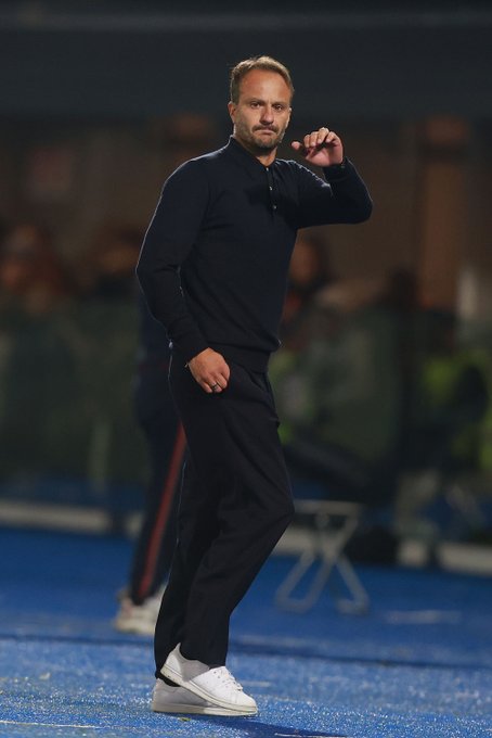 A male coach with short blond hair and a focused expression wears a black sweater and black pants standing on a blue-turfed soccer field gesturing emphatically with his right hand raised palm forward while his left hand rests in his pocket. Blurred spectators and team members in dark clothing appear in the background under stadium lighting. White sneakers are visible on his feet. The setting suggests a professional match environment.
